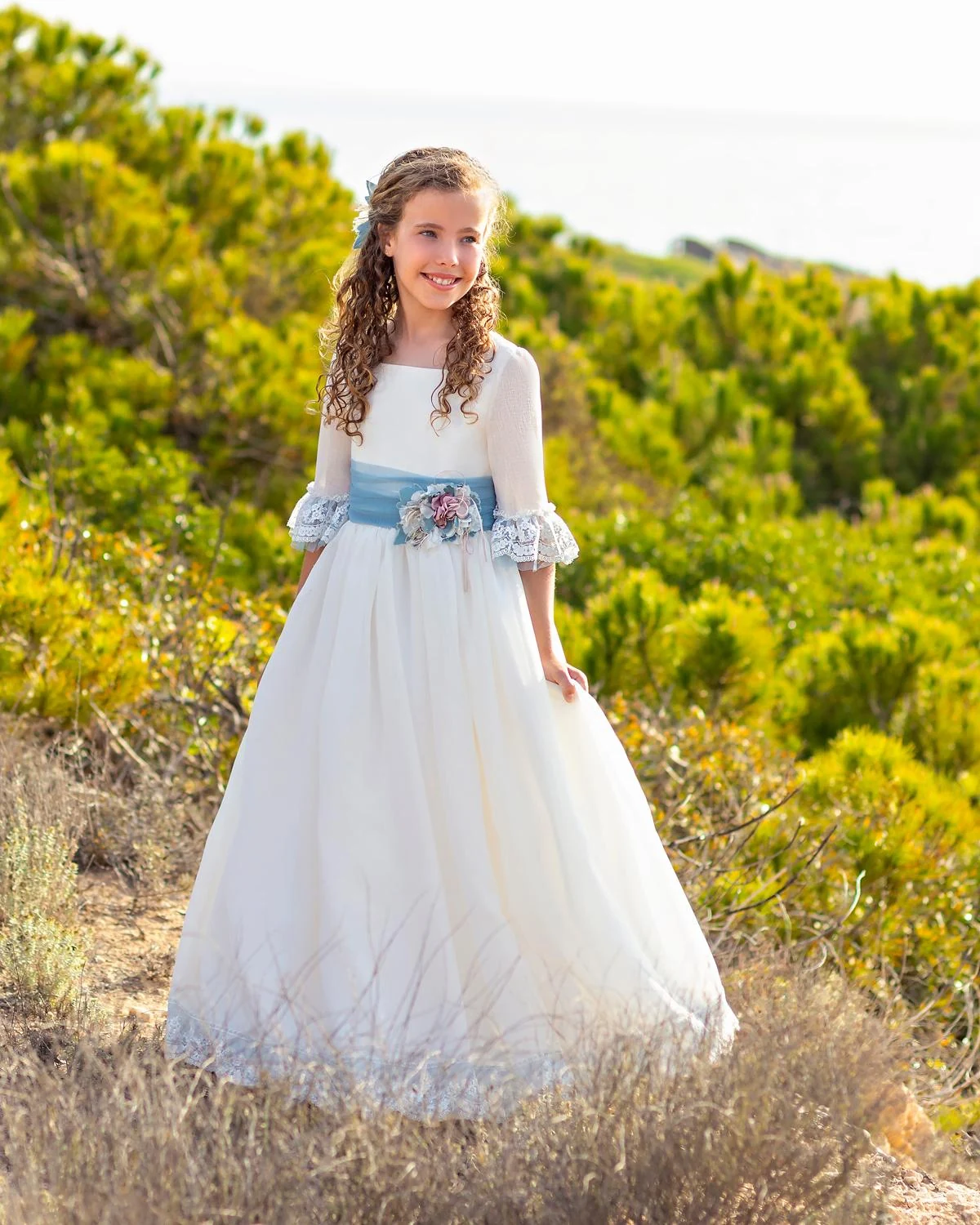 Vestido de comunión Carmy 5804 en exterior, de frente, con fajín de tul azul y flores en la cintura